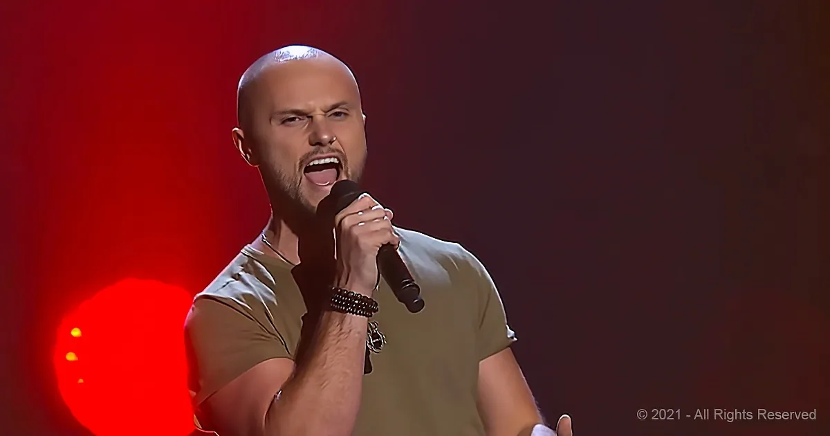 A man with a shaved head and beard sings passionately into a microphone, wearing a bracelet, against a red-lit background.