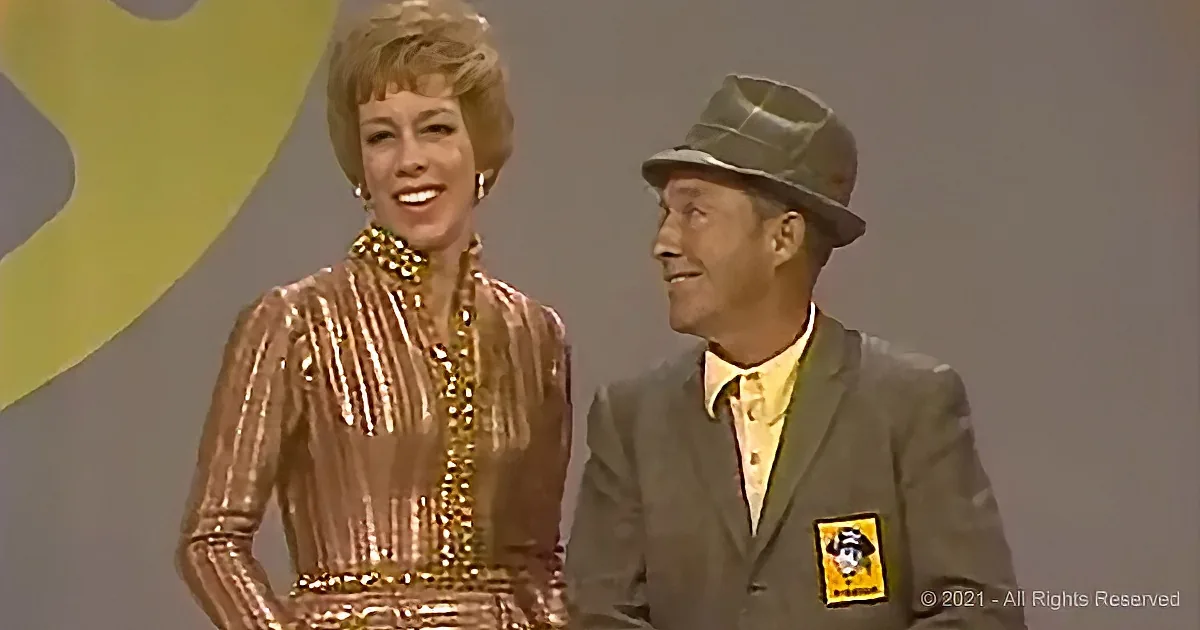 A woman in a shimmering dress and a man in a suit with a hat are smiling and standing together on a stage.
