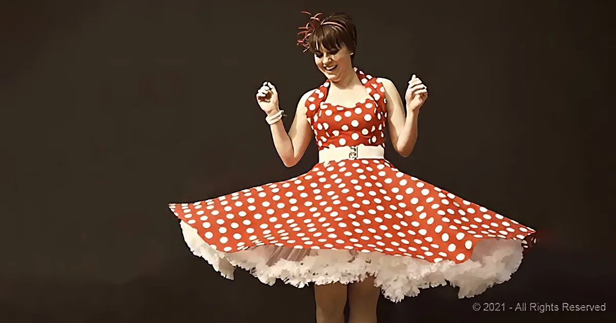 A woman in a red polka-dot dress with a white belt and white frills twirls joyfully against a dark background.