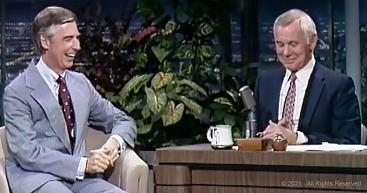 Two men in suits sit at a table, one laughing, in a studio setting with plants and city lights in the background.