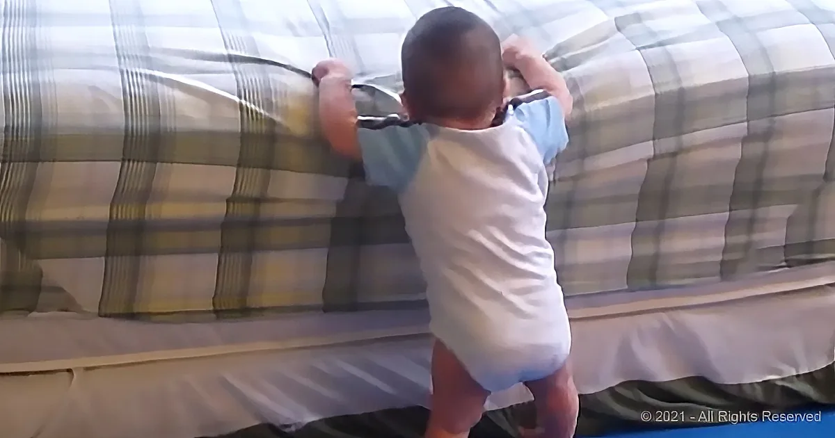 A baby in a white onesie is climbing a plaid-patterned couch.