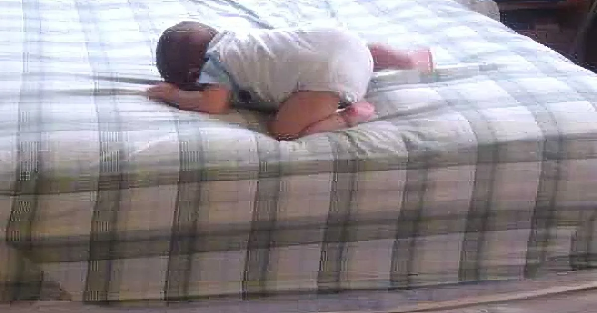 A baby in a white onesie is climbing a plaid-patterned couch.