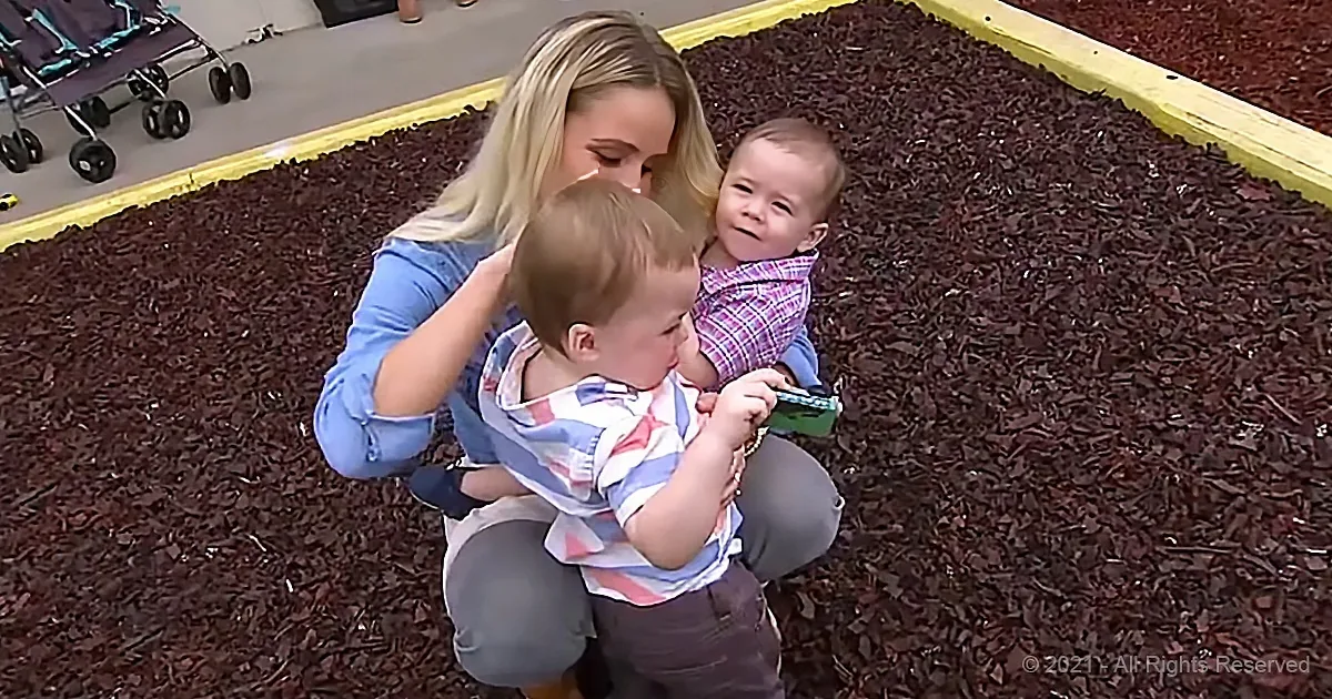 A woman holds two young children in a mulched area, with a stroller in the background.