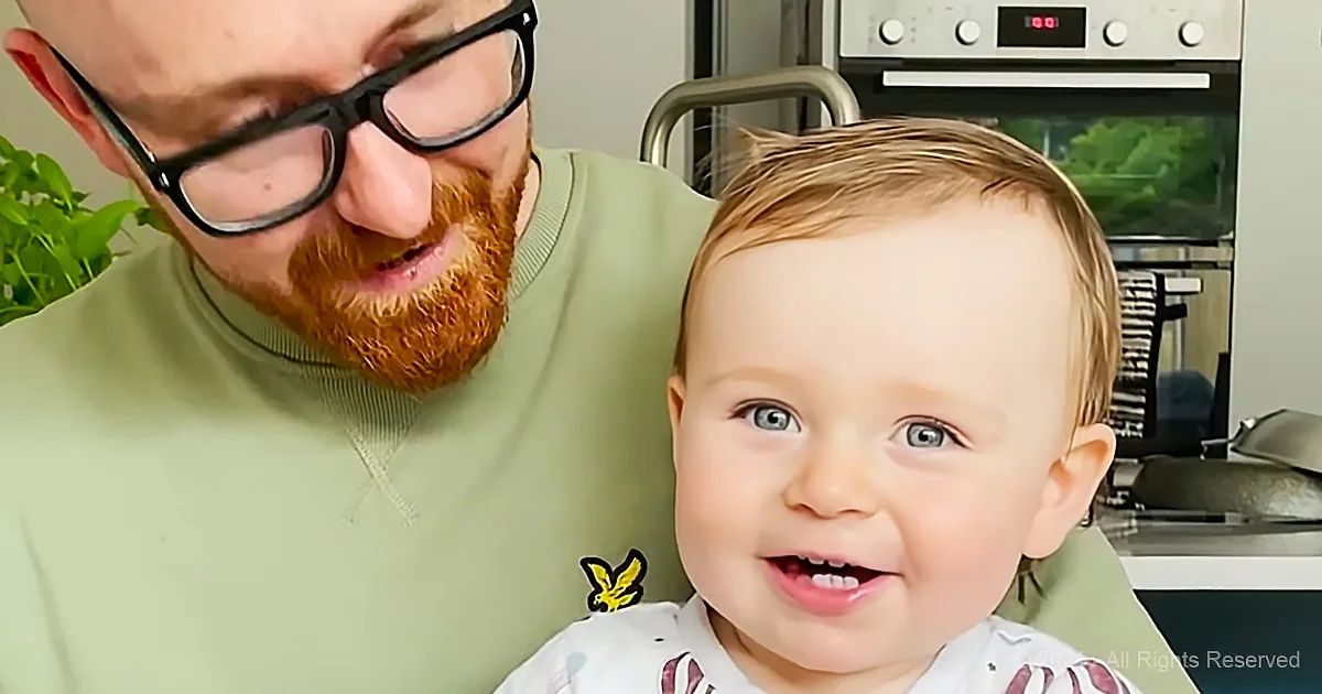 A smiling baby with blue eyes and a man with glasses and a beard in a kitchen.