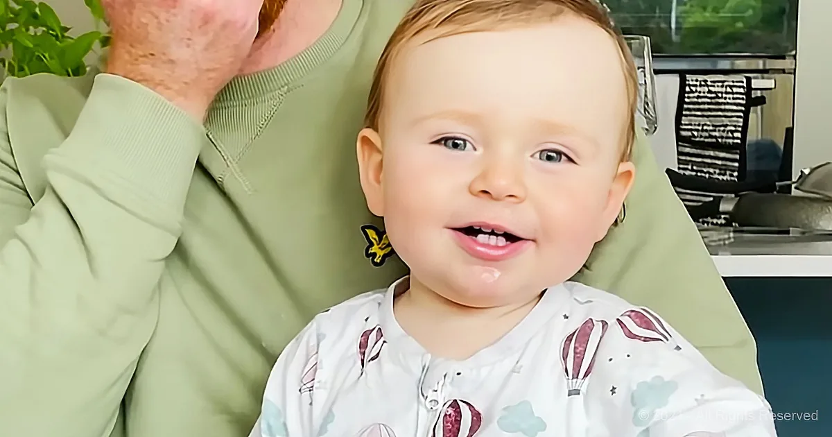 A smiling baby in a white shirt with hot air balloons, held by a person in a green shirt.