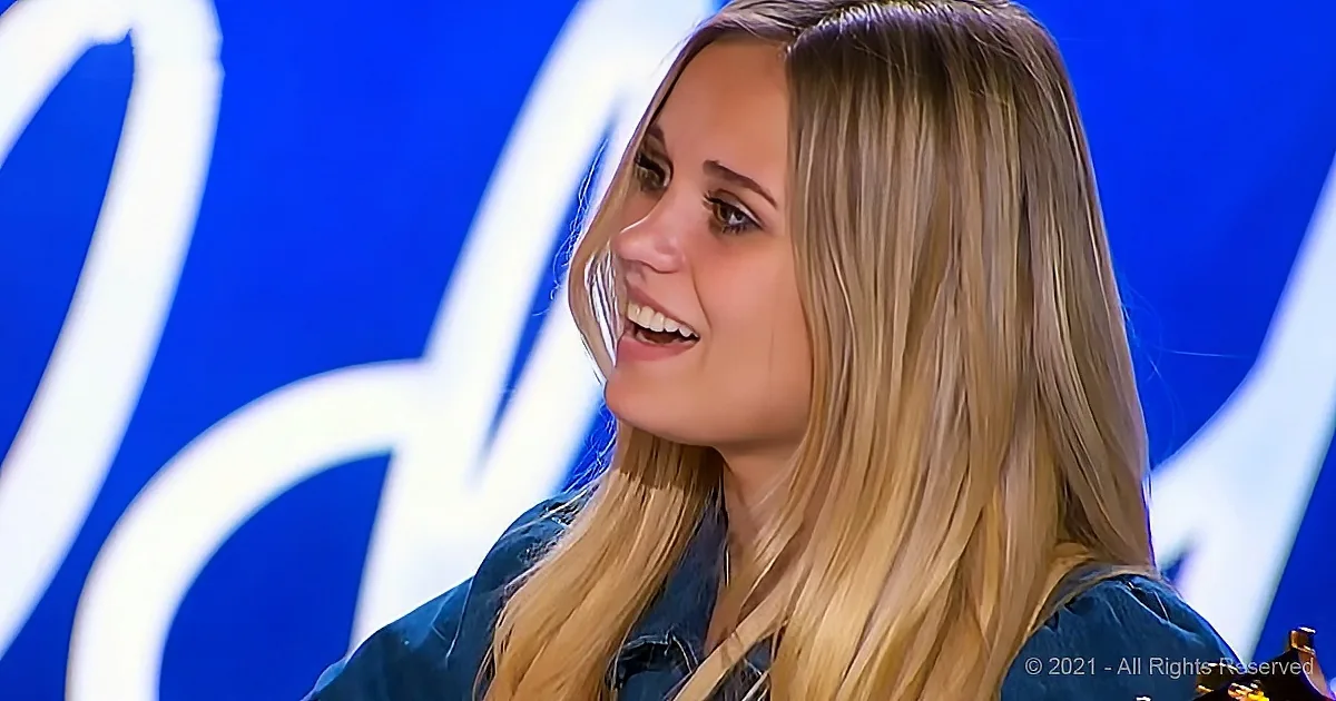 A woman with long blonde hair smiles, wearing a denim jacket, in front of a blue background with white lettering.