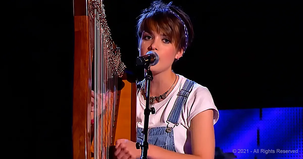 A person wearing overalls sings into a microphone while playing a harp.