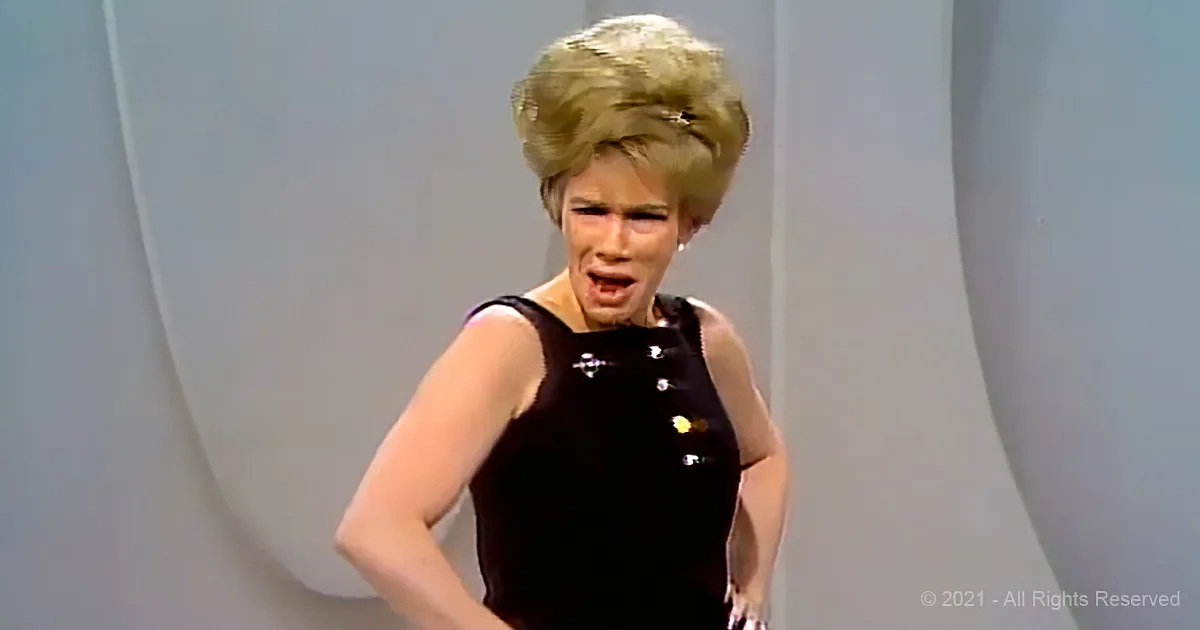 A person in a black dress with a large, styled blonde wig is standing with hands on hips.