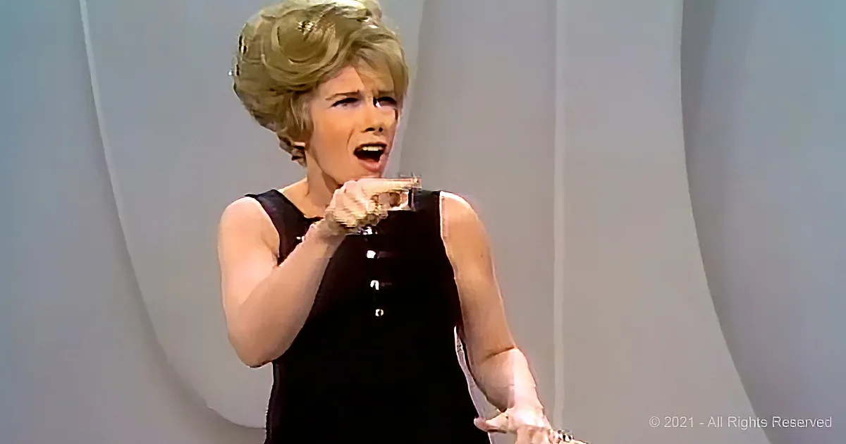 A woman in a black dress is holding a small object and appears to be speaking or singing.