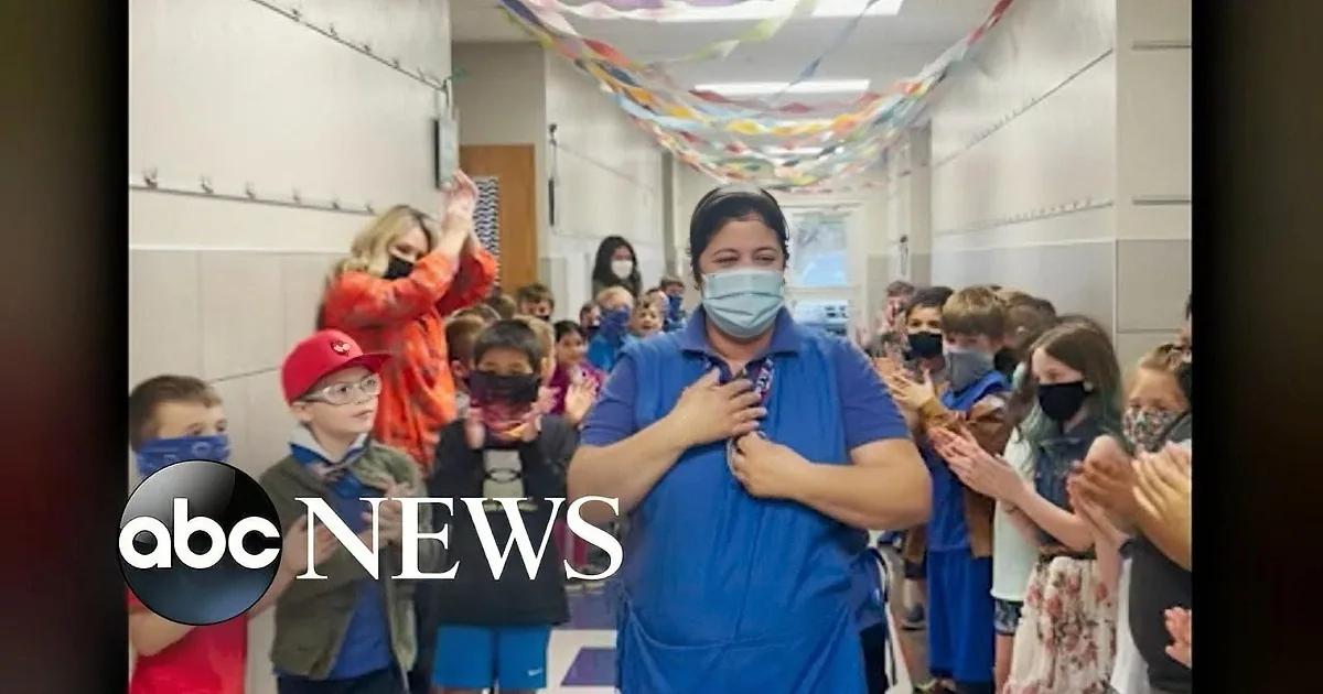 A woman in a blue shirt holds a microphone, surrounded by children clapping and wearing masks.