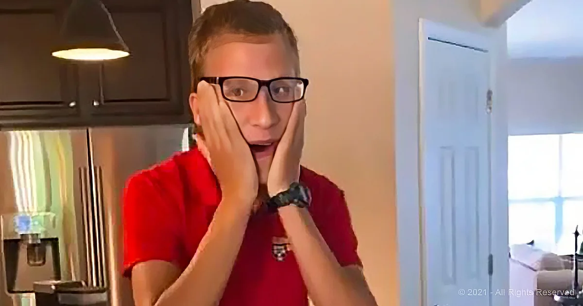 A person in a red shirt covers their mouth with both hands, wearing glasses and a watch, in a kitchen setting.