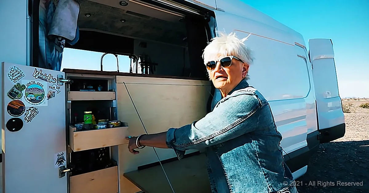 A woman in sunglasses opens a cabinet in a van, revealing a small kitchenette with food items.