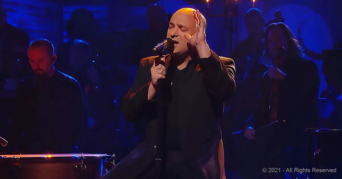 A man in black holds a microphone, singing with his hand on his head, surrounded by musicians in a dimly lit stage setting.