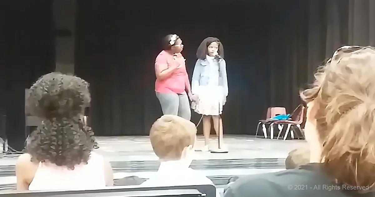 Two women stand on stage, one in pink, the other in blue, facing each other, with audience in foreground.
