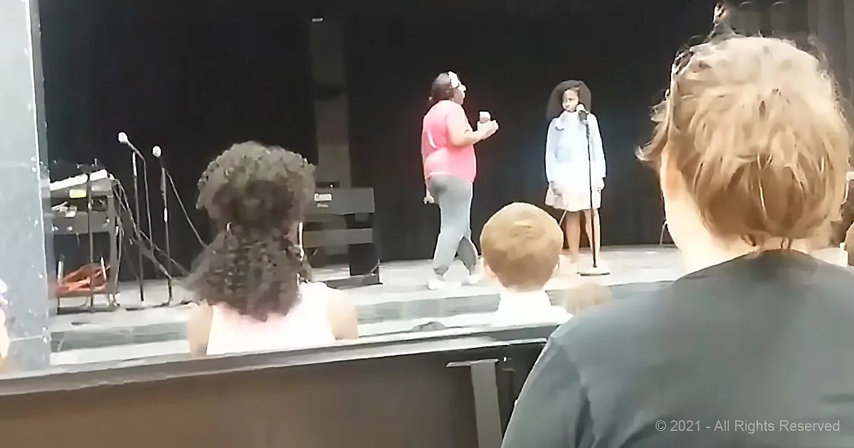 A woman in a pink shirt stands on stage with a microphone, addressing an audience.
