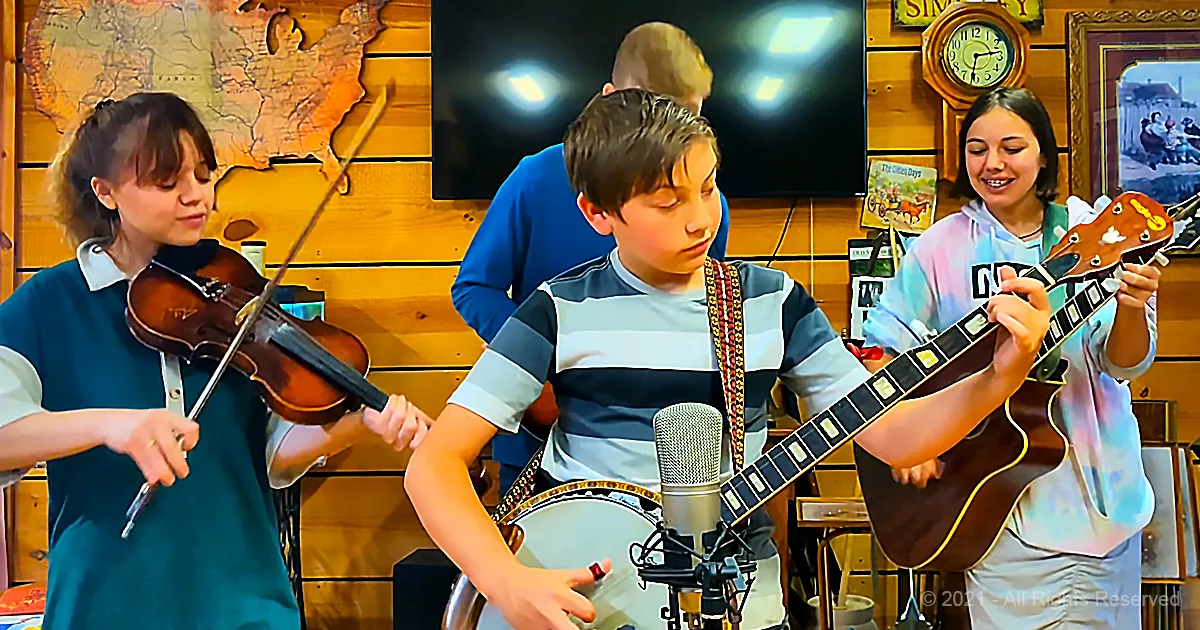 A young boy plays banjo in front of a microphone, surrounded by other musicians and a wooden wall with a map and clock.