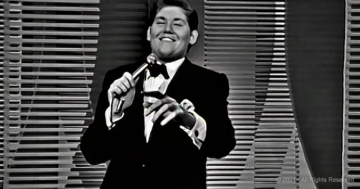 A man in a tuxedo smiles while holding a microphone, standing against a striped backdrop.