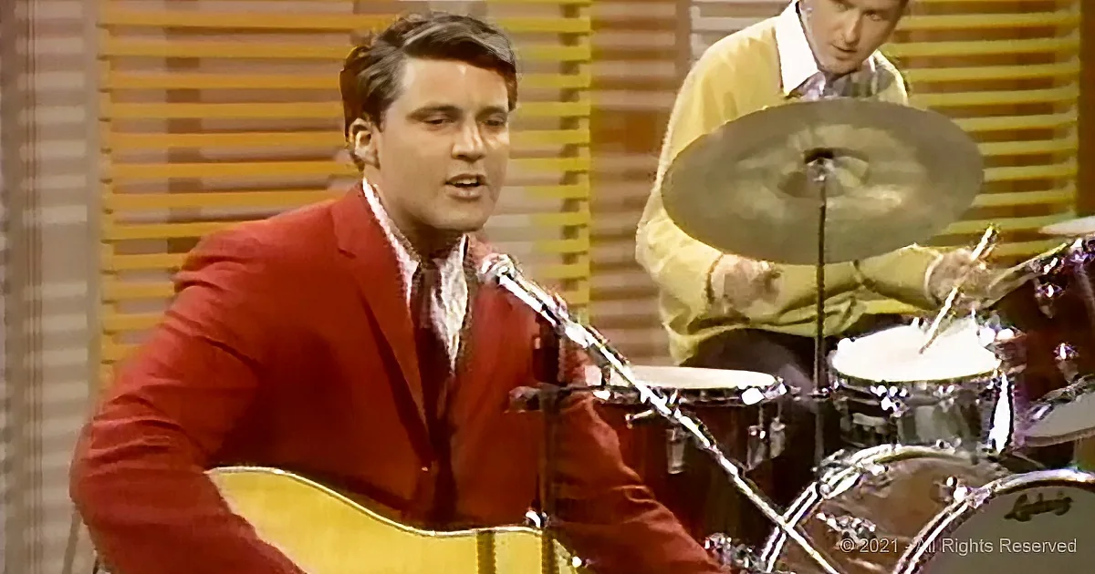 A man in a red suit plays guitar while singing, with a drummer in the background.