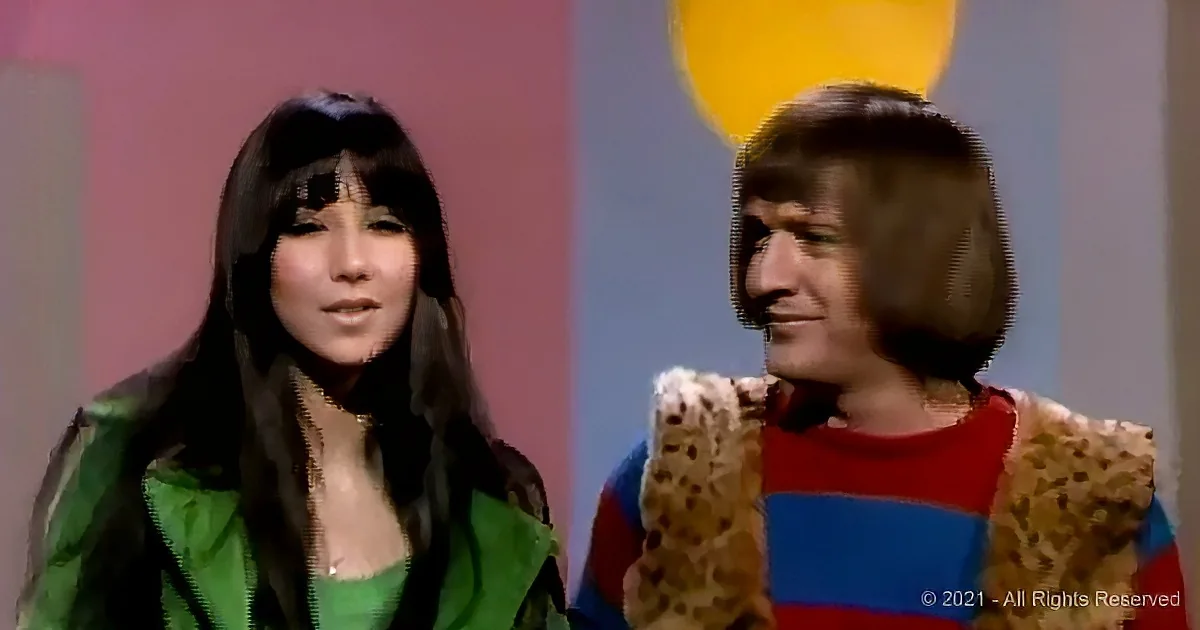 A woman in a green jacket and a man in a red and blue sweater with a fur vest are standing side by side.