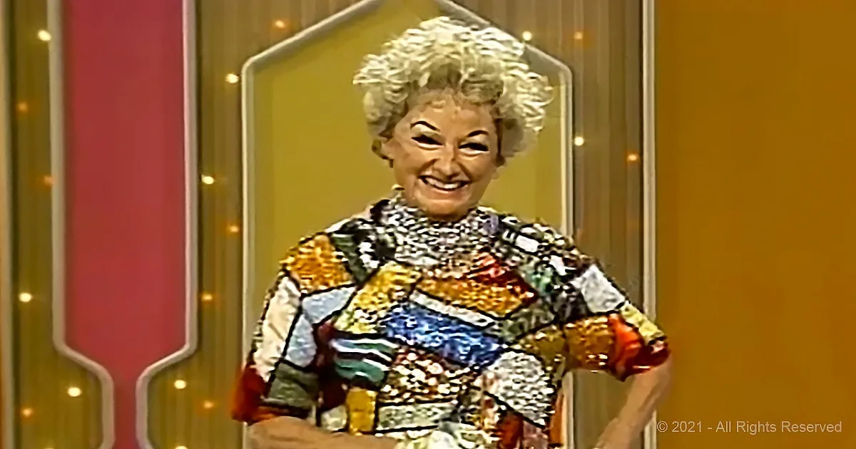 A smiling woman with curly hair, wearing a colorful, patterned dress, stands in front of a decorative background.
