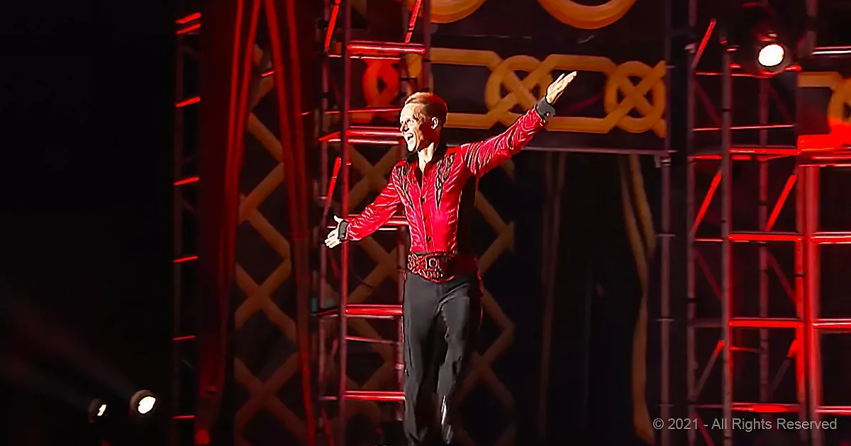 A man in a red shirt and black pants stands on stage with arms outstretched, illuminated by red lighting.