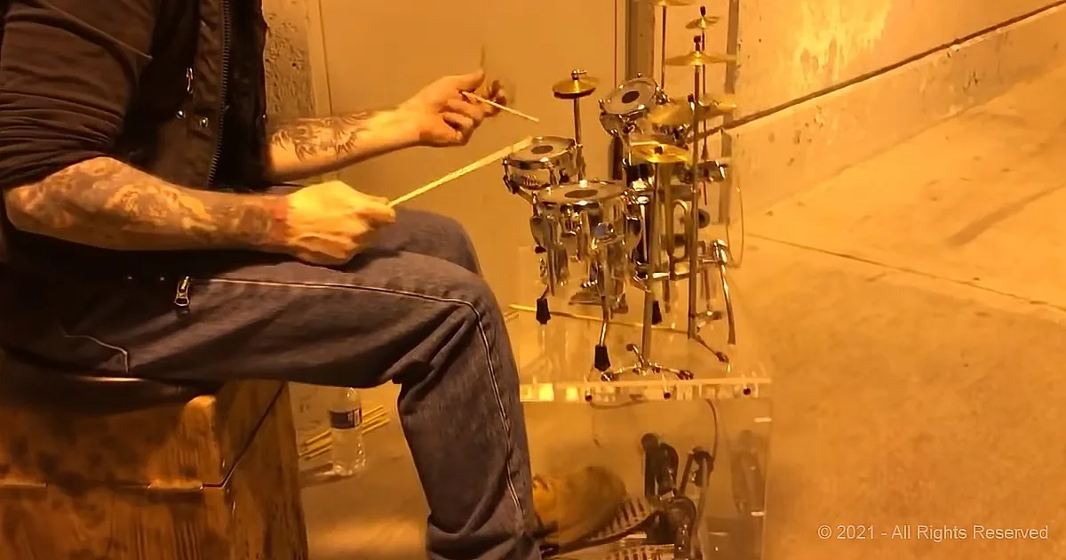 A tattooed person sits on a stool, holding drumsticks, with a small drum set in front of them.