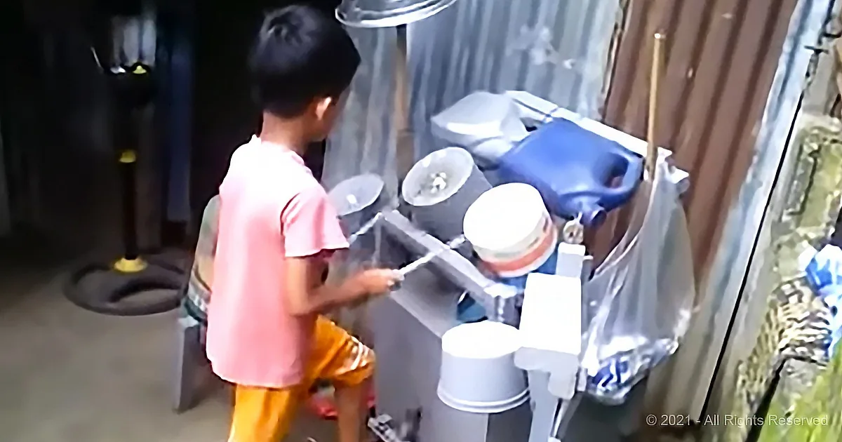 A child in a pink shirt and yellow shorts is washing dishes at a sink with buckets and a hose.