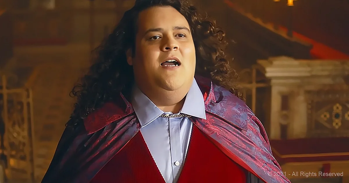 A person with curly hair, wearing a red cape and blue shirt, stands in a dimly lit, ornate setting.