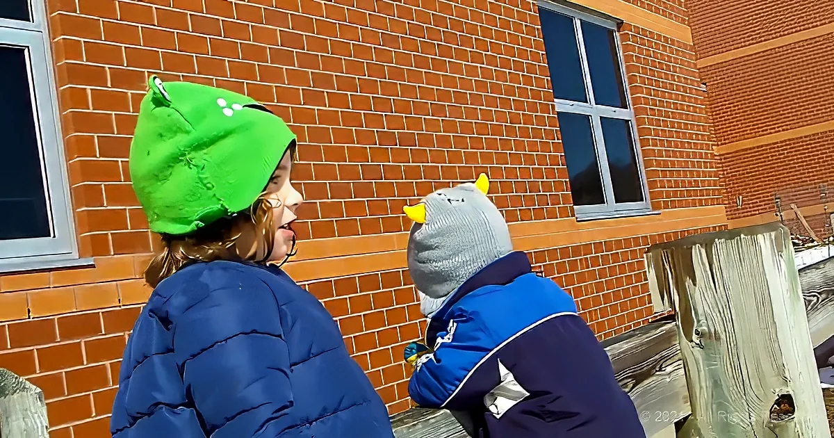 Two children in winter coats and hats stand near a brick building, one with horns.