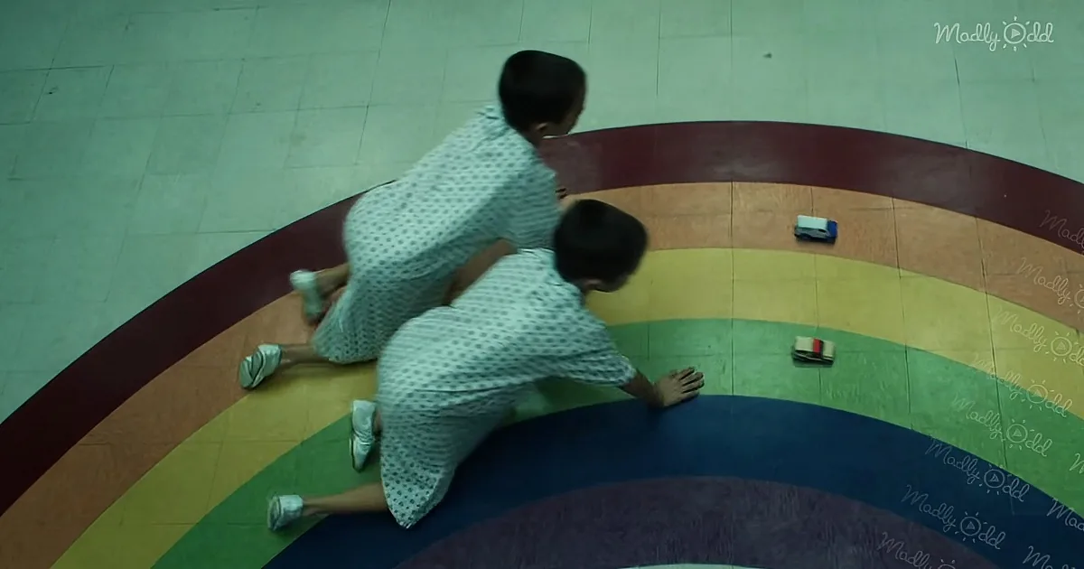 Two children in polka-dot dresses crawl on a colorful rainbow-patterned floor, reaching for toy cars.