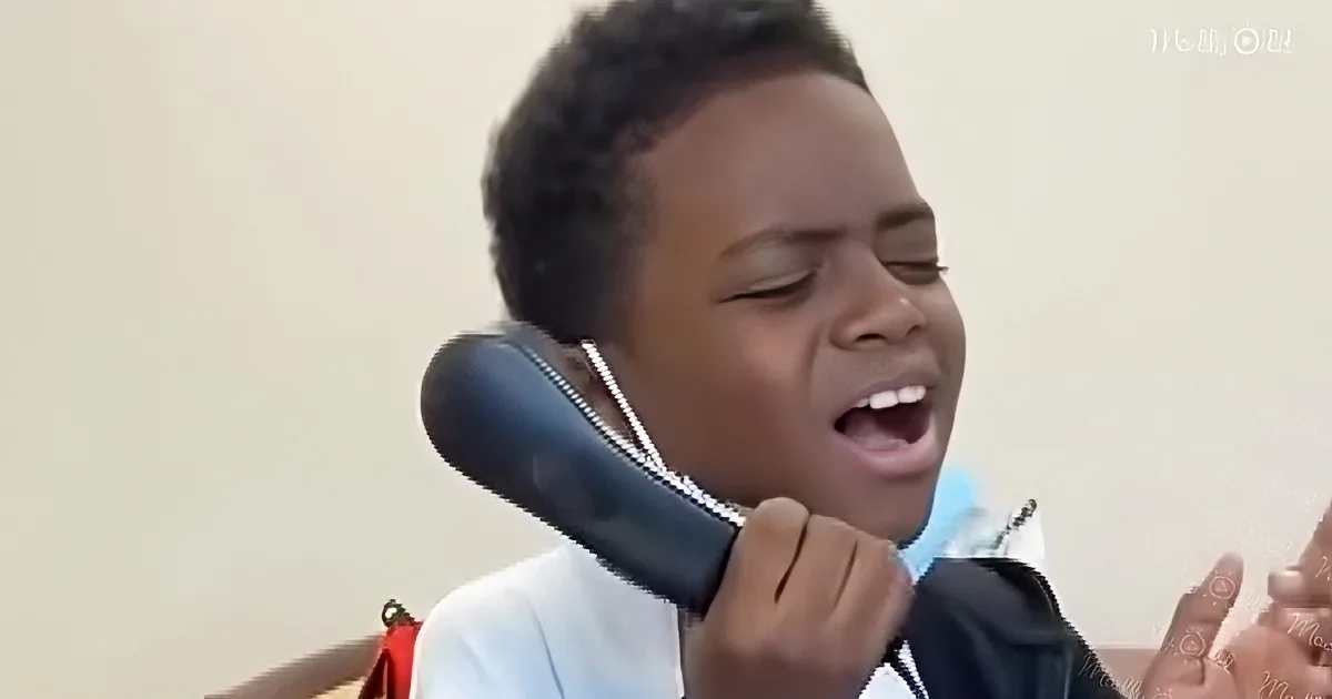 A young boy with short hair is holding a black object to his ear, appearing to talk or sing.
