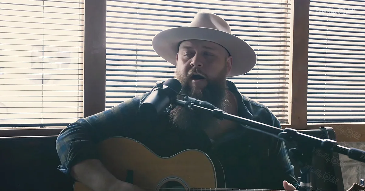 A man with a beard and hat sings into a microphone while playing an acoustic guitar.