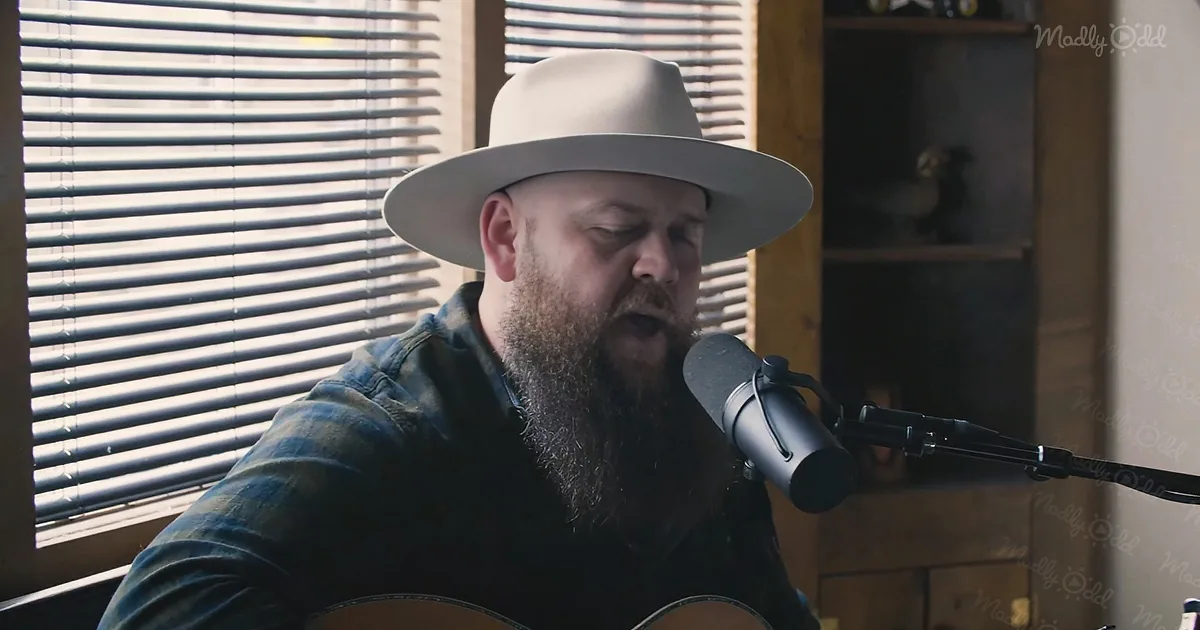 A man with a beard and a wide-brimmed hat sings into a microphone while playing guitar.