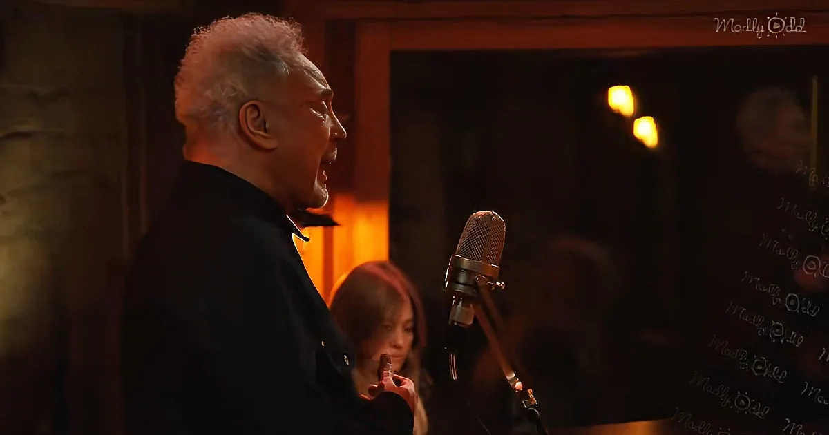 A man sings into a microphone, illuminated by warm lighting, with another person in the background.