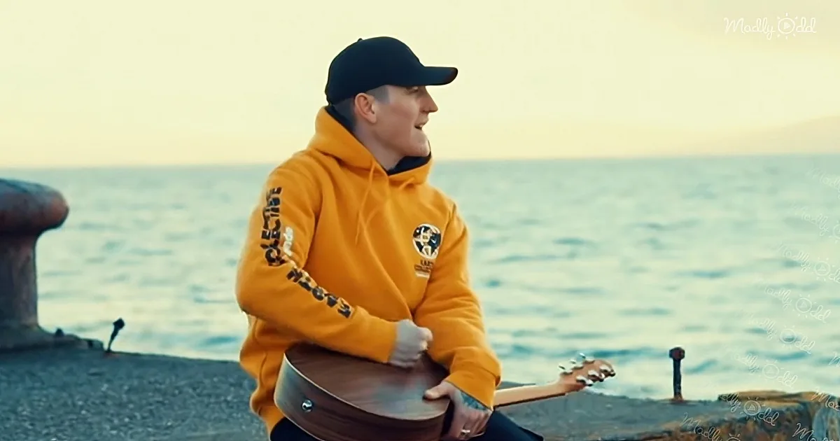 A person in a yellow hoodie and black cap plays guitar by the sea at sunset.