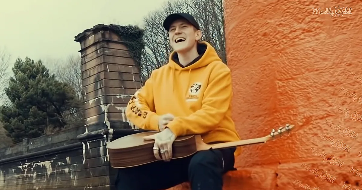 A person in a yellow hoodie and cap sits against a red wall, holding a guitar, smiling.