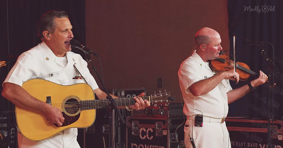 A man in white uniform sings and plays guitar on stage, accompanied by another man playing violin.