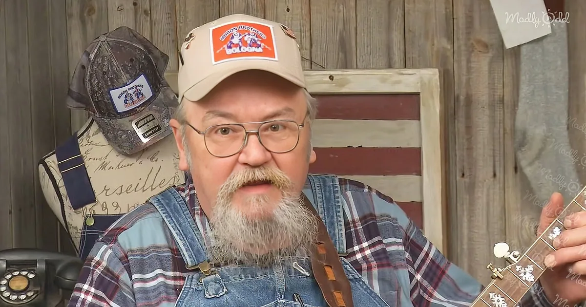 A man with a beard and glasses wearing overalls and a hat with "Bologna" patch, holding a guitar.