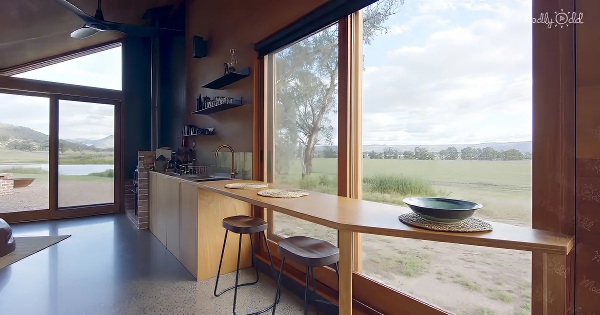 A modern kitchen with a long wooden counter, stools, and a view of a grassy field and distant hills.