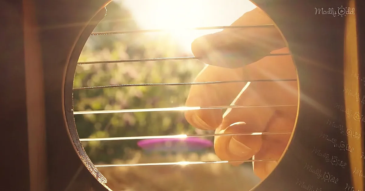A hand plucks guitar strings with sunlight streaming through, creating a warm, serene atmosphere.