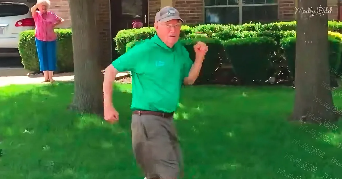A man in a green shirt and cap dances joyfully on a grassy lawn, with a woman in the background.