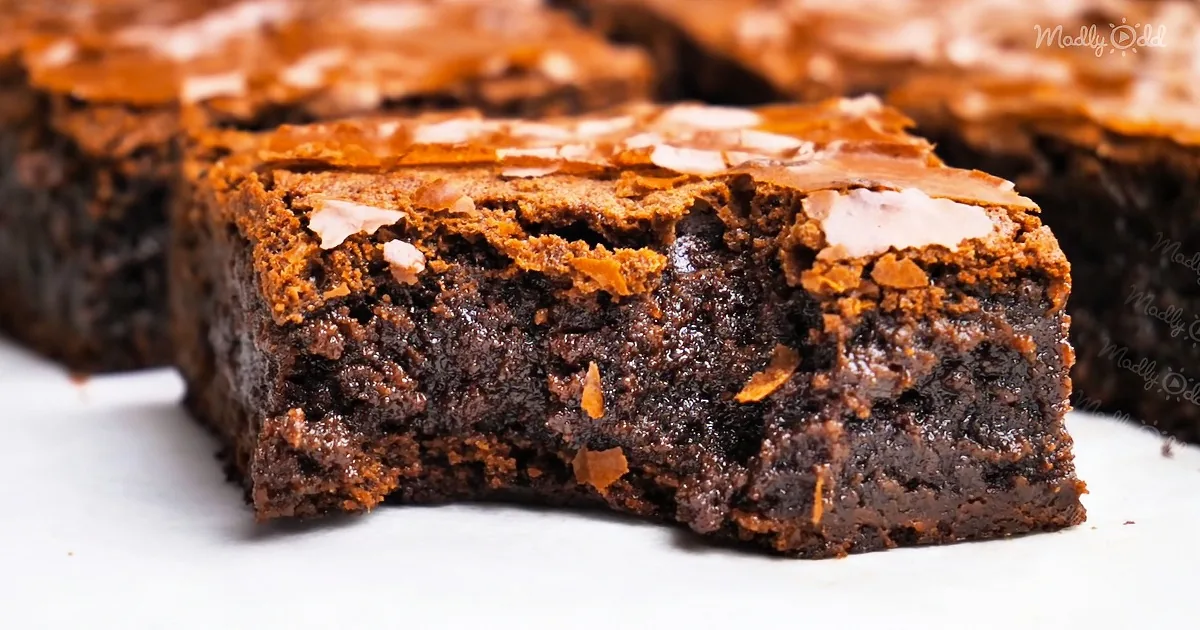 A rich, chocolatey brownie with a glossy surface and crumbly edges, topped with chocolate shavings.