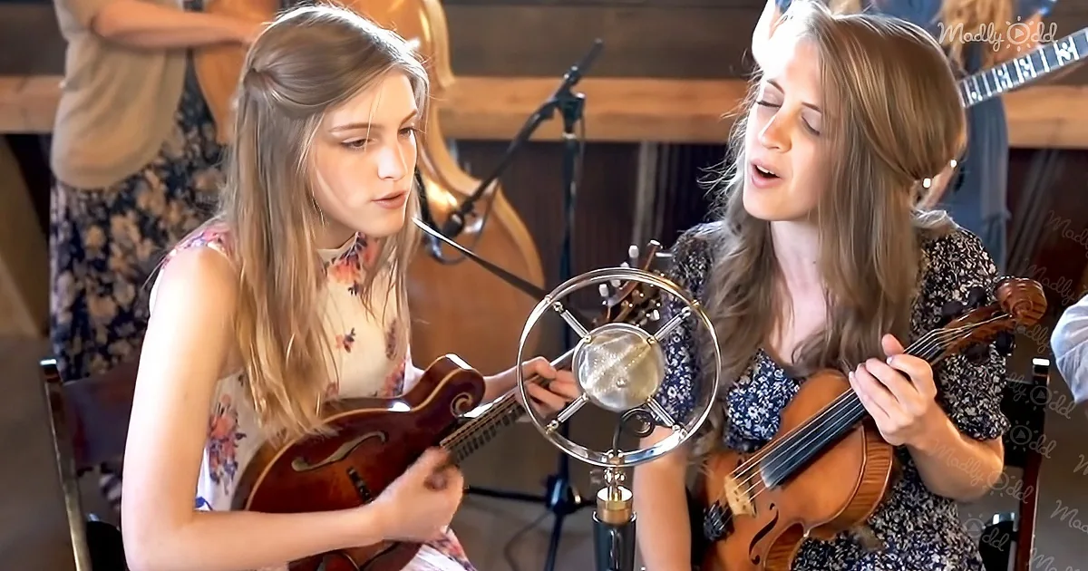 Two women play mandolin and violin in front of a microphone, singing together.