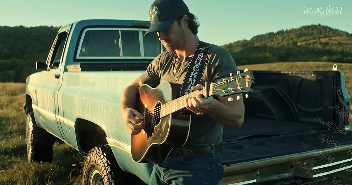 A man in a cap plays an acoustic guitar in front of a vintage pickup truck in a scenic, sunlit field.