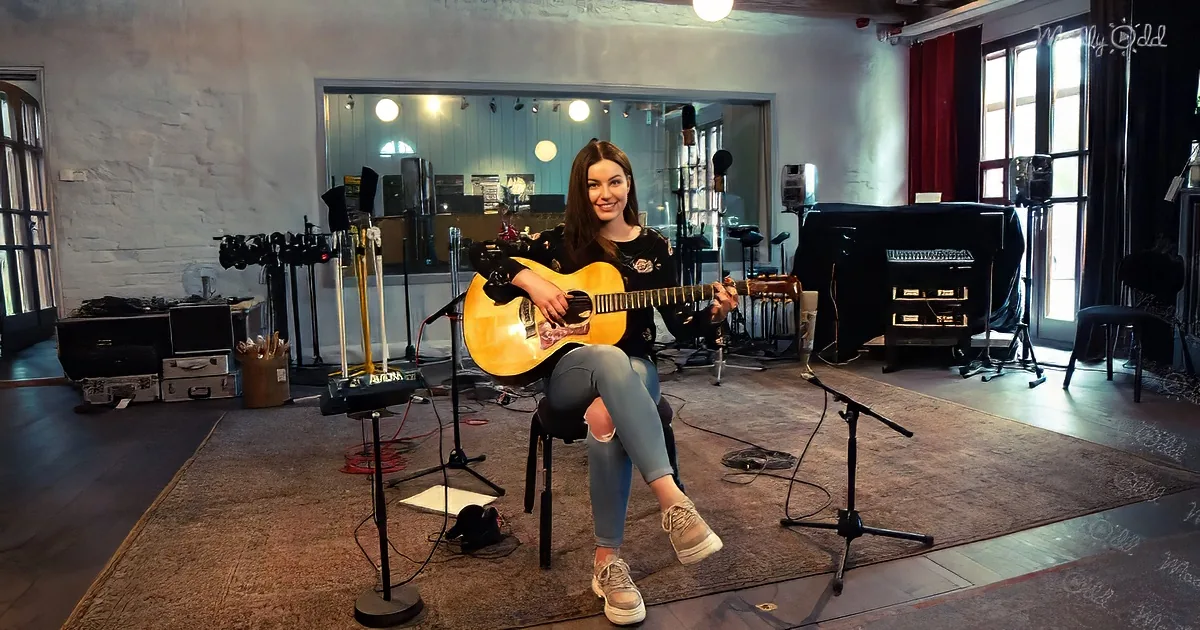 A woman sits on a stool in a recording studio, holding an acoustic guitar and smiling.
