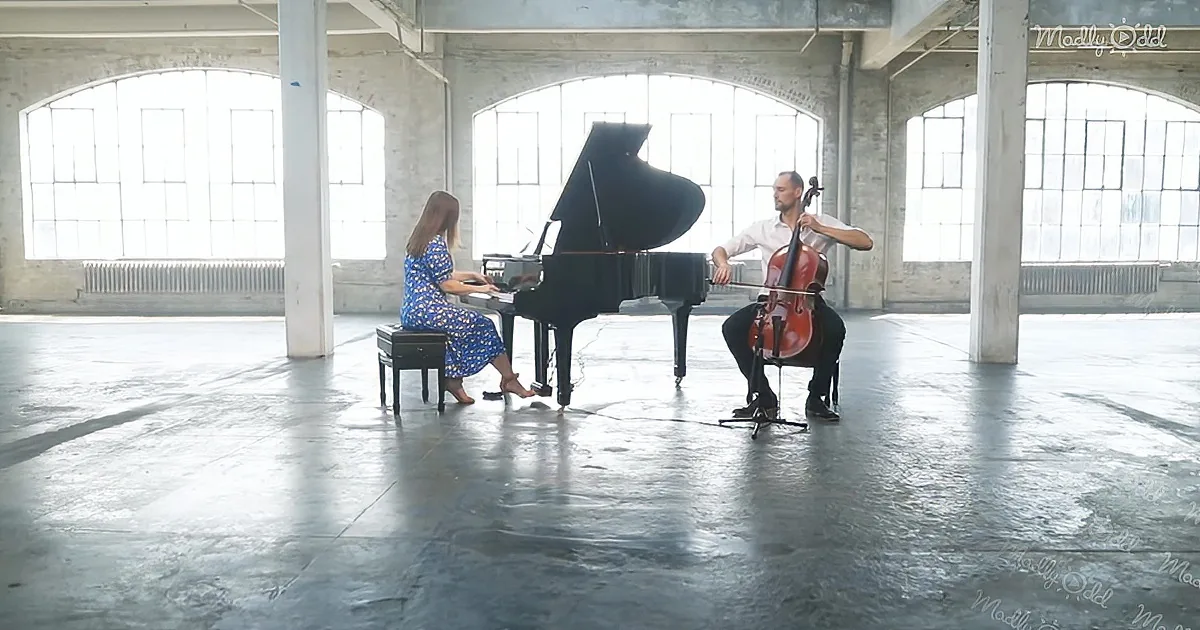 A woman plays piano while a man plays cello in a spacious, industrial-style room with large windows.