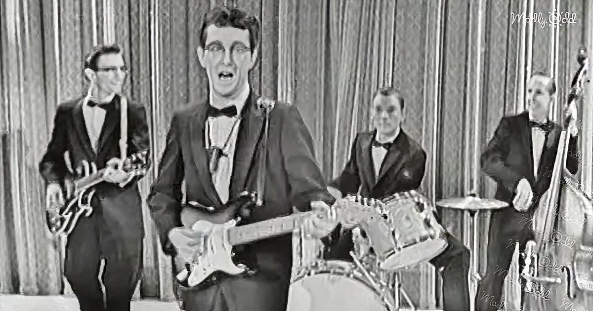 A man in a suit plays guitar while singing, with a band behind him.