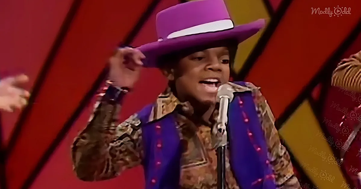 A person wearing a purple hat and colorful outfit sings into a microphone on a stage with a red background.