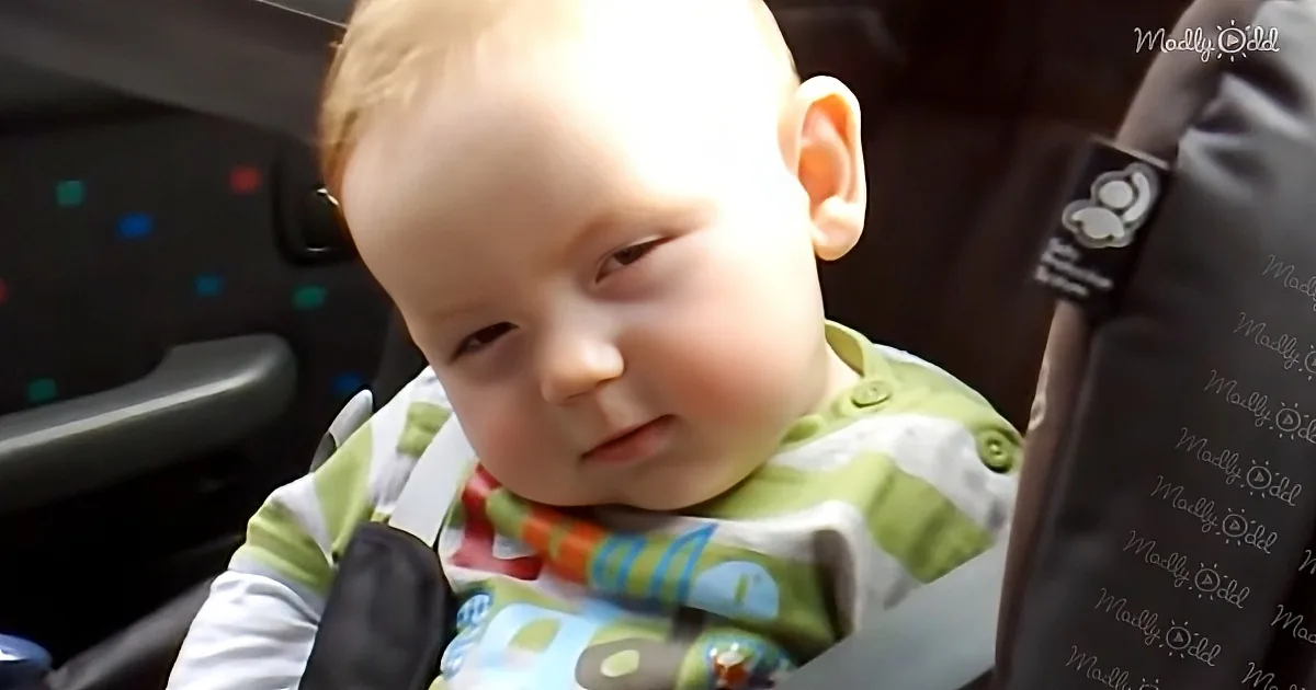 A baby in a car seat wearing a green and white outfit, looking down with a curious expression.