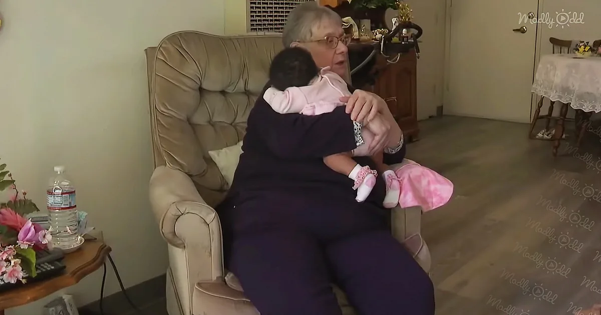 An elderly woman sits in a chair, holding a baby dressed in pink, surrounded by a cozy living room setting.